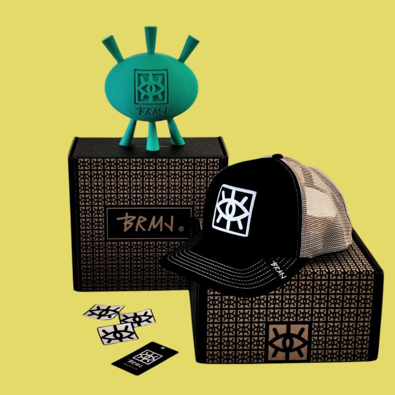 Caja negra con una gorra y una escultura verde con el logo de brmjcaps sobre fondo amarillo.