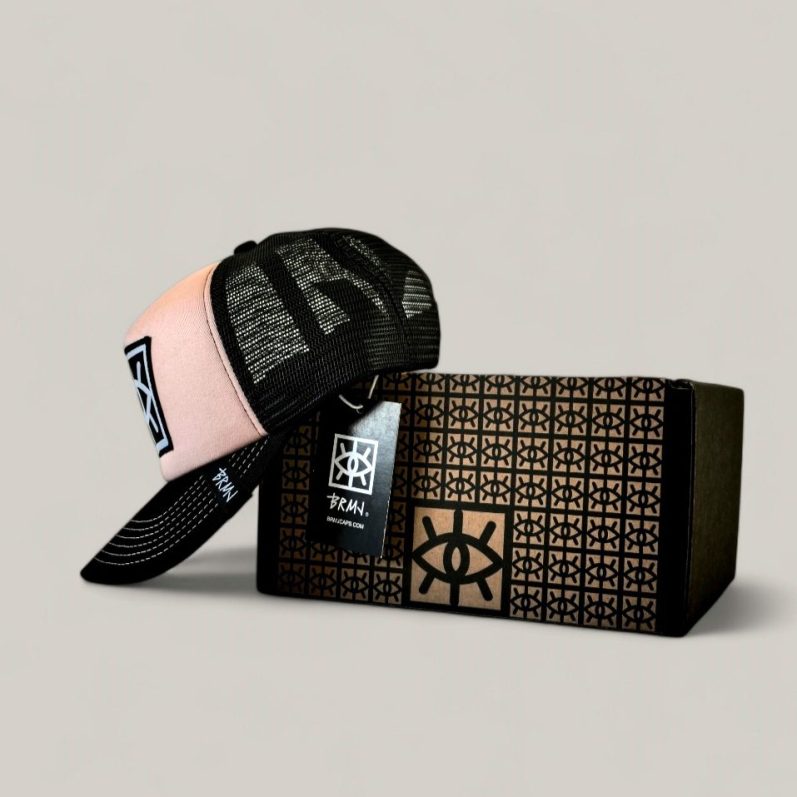 Gorra negra con visera rosa apoyada sobre una caja con diseño gráfico de un ojo y logotipo de la marca brmjcaps